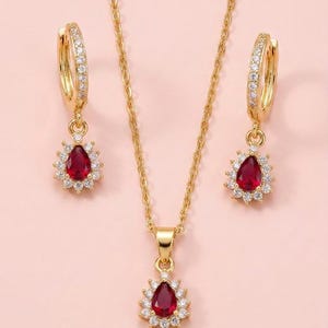 May include: A gold-toned jewellery set consisting of a necklace and two hoop earrings. Each piece features a central pear-shaped red stone surrounded by a halo of small clear stones. The necklace has a delicate chain and a matching pendant.