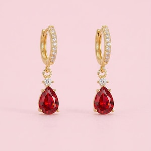 May include: A pair of gold hoop earrings featuring a row of small clear stones along the hoop. Each earring has a dangling charm consisting of a single small clear stone above a larger, pear-shaped, deep red faceted gemstone in a gold setting.
