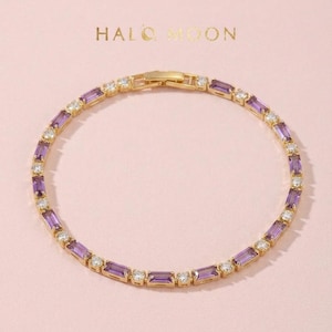 May include: A gold-toned bracelet featuring alternating baguette-cut purple stones and round clear stones. The jewellery piece is arranged in a circular shape with a secure clasp mechanism. The text HALO MOON is printed in gold above the bracelet.