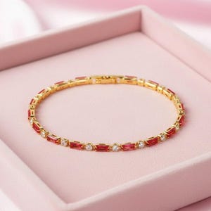 May include: A gold bracelet featuring alternating red and clear rectangular stones. The jewellery is displayed on a pink surface. The clasp is visible. The design highlights the contrast of colours.