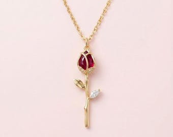 May include: A gold-tone necklace featuring a pendant shaped like a rosebud with a deep red faceted stone at the center, accented by small clear crystals at the base, a gold stem, two gold leaves, and one small clear marquise-cut crystal leaf.