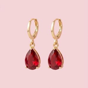 May include: A pair of gold-tone hoop earrings featuring dangling teardrop-shaped red gemstones. Each earring consists of a small circular hoop connected to a faceted, deep red pear-cut stone held in a gold-coloured metal setting with prongs.