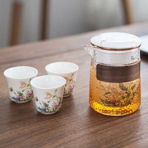 May include: A tea set comprising a glass teapot with a white lid and a wooden band, and three small teacups. The teapot has a textured design and contains a golden liquid. The teacups are white with floral patterns.