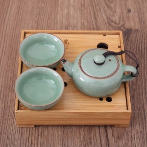 May include: A celadon green ceramic tea set, comprising a teapot and two small cups, displayed on a bamboo tray. The teapot features a matching lid and a dark brown trim. The cups and teapot have a crackle glaze finish.