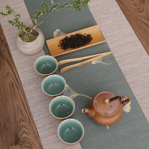 May include: A tea set arranged on a patterned runner. The set includes a brown teapot, four teal and brown teacups, a bamboo tray with tea leaves, and a small bonsai tree in a ceramic pot. Wooden tongs are also included.
