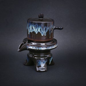 May include: A black ceramic teapot with a blue and green glaze sits on a three-legged stand. The teapot has a lid and a spout.