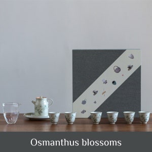 May include: A tea set featuring a teapot, six small cups, a glass pitcher, and a gift box. The teapot and cups are decorated with a floral pattern. The grey gift box has a diagonal white stripe and decorative images. The text "Osmanthus blossoms" is at the bottom.