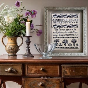 May include: A wooden table displays a framed cross-stitch sampler with blue embroidery and text. Beside it are a vase of flowers, a lit candle, and a glass bowl. The sampler reads "The flowers appear on the earth, the time for singing has come."