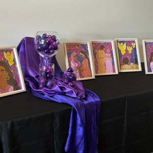May include: A display of framed art prints with religious themes, alongside decorative purple baubles in glass containers. The artwork depicts figures with stained-glass style backgrounds. The display is set on a black tablecloth with a purple satin drape.