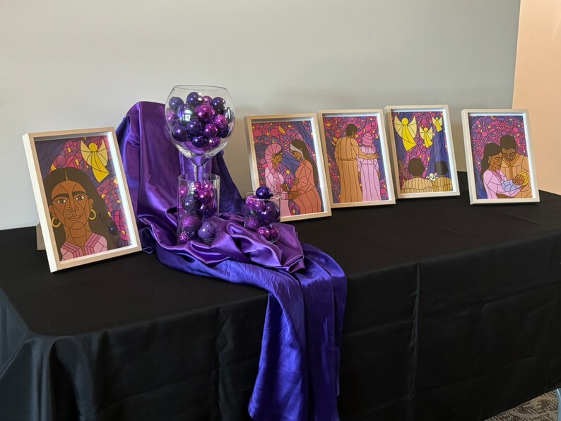 May include: A display of framed art prints with religious themes, alongside decorative purple baubles in glass containers. The artwork depicts figures with stained-glass style backgrounds. The display is set on a black tablecloth with a purple satin drape.