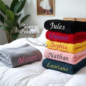 Op de afbeelding: Een stapel van zes fleece dekens in verschillende kleuren, elk met een andere naam geborduurd in een contrasterende kleur. De namen zijn Jules, Clement, Sophia, Nathan, Lauriane en Manon.