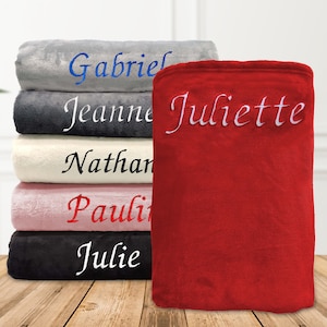 Peut inclure: Une pile de six couvertures en peluche de différentes couleurs, chacune avec un nom différent brodé en fil blanc. Les noms sont Gabriel, Jeanne, Nathan, Pauli, Julie et Juliette. La couverture du dessus est rouge et porte le nom Juliette brodé.