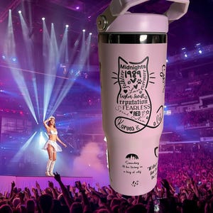 May include: A light pink insulated tumbler with a white handle and lid. The tumbler features a black cat graphic with text including "Midnights 1989", "Karma is a cat", and other song titles. The background shows a concert stage.