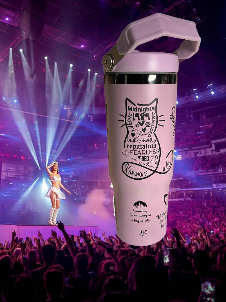 May include: A light pink insulated tumbler with a white handle and lid. The tumbler features a black cat graphic with text including "Midnights 1989", "Karma is a cat", and other song titles. The background shows a concert stage.