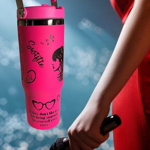 May include: A bright pink tumbler with a matching handle and lid. The tumbler features black graphics, including a silhouette of a woman, musical notes, and the word "Swiftie." The text "If they don't like you for being yourself, be yourself even more" is also printed on the tumbler.