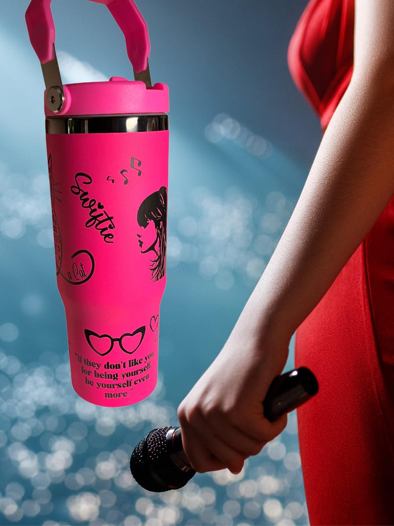 May include: A bright pink tumbler with a matching handle and lid. The tumbler features black graphics, including a silhouette of a woman, musical notes, and the word "Swiftie." The text "If they don't like you for being yourself, be yourself even more" is also printed on the tumbler.