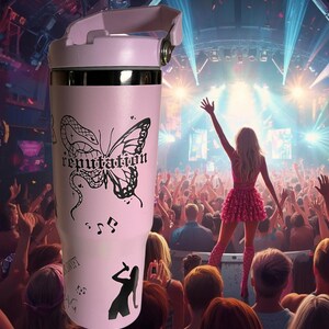 May include: A light pink tumbler with a black butterfly and the word "reputation" printed on it. The tumbler also features musical notes and a silhouette of a singer. The background shows a concert stage.