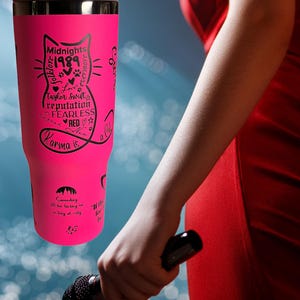 May include: A bright pink tumbler featuring a black cat design with song titles and the text "Midnights 1989 folklore &evermore Love Taylor Swift reputation FEARLESS RED Karma is". The tumbler has a handle and a silver lid.