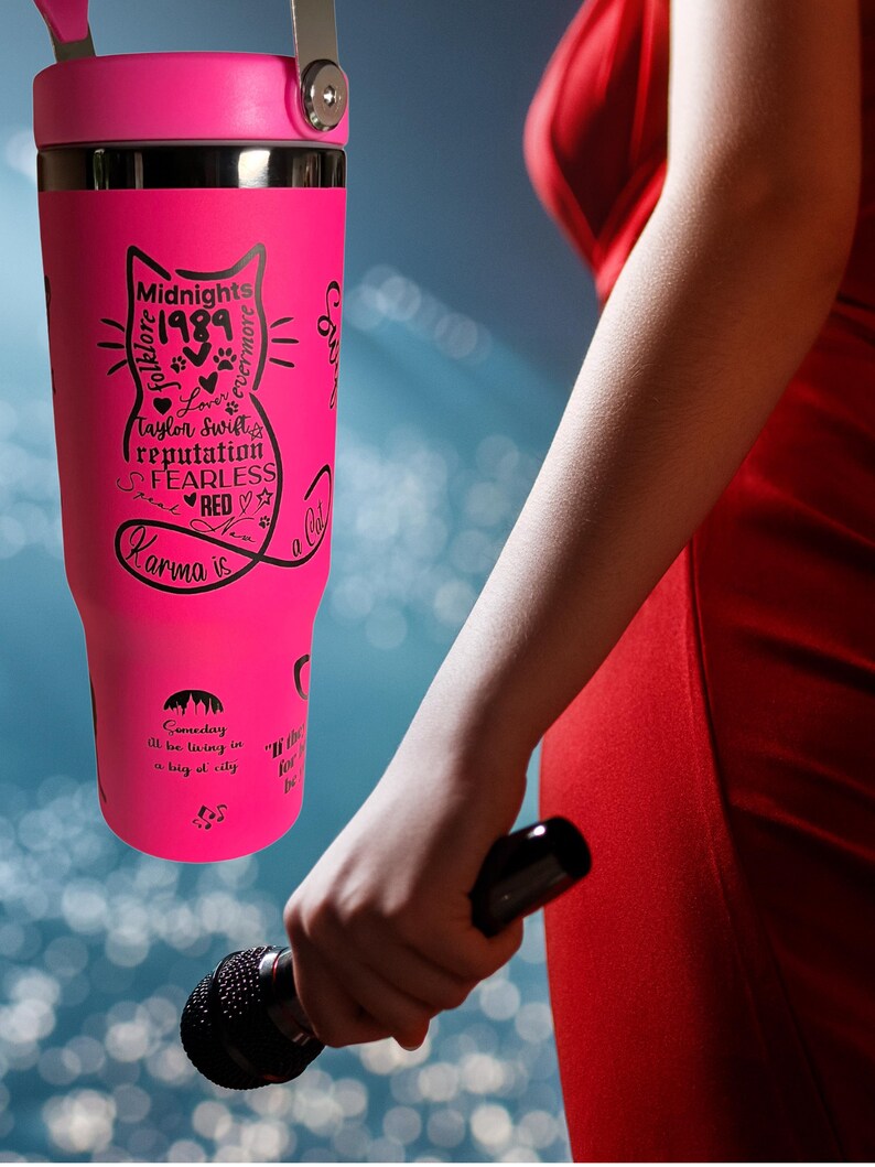 May include: A bright pink tumbler featuring a black cat design with song titles and the text "Midnights 1989 folklore &evermore Love Taylor Swift reputation FEARLESS RED Karma is". The tumbler has a handle and a silver lid.