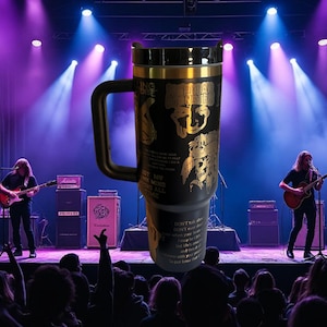 Może przedstawiać: Czarny i złoty kubek podróżny z uchwytem, ​​z motywem zespołu. Kubek ma czarny korpus ze złotą grafiką i tekstem. Tło przedstawia występ muzyczny na żywo z muzykami na scenie i publicznością.