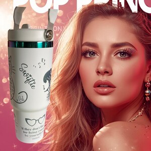 May include: White tumbler with a handle and a lid. The tumbler has a green band and a design that includes a cat, musical notes, and the word "Swiftie". The tumbler also has the quote "If they don't like you for being yourself, be yourself more."