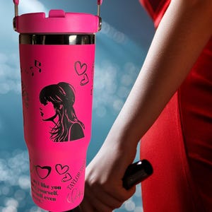 May include: A pink insulated tumbler with a handle, featuring a black silhouette of a woman, musical notes, and hearts. The tumbler has the text "Taylor Swift" and a quote. The tumbler is held by a person wearing a red dress.