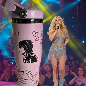 May include: A light pink insulated tumbler with a black and white silhouette of a woman, musical notes, and hearts. The tumbler has the text "Taylor Swift" and a signature. The lid is light purple.