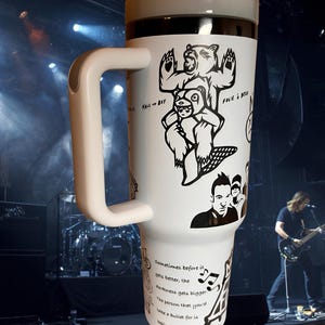 Könnte beinhalten: Weißer Reisebecher mit weißem Griff und Deckel. Der Becher zeigt schwarze Illustrationen von Bären und Text, darunter "Fall Out Boy" und Songtexte. Der Hintergrund ist eine Konzertbühne mit Musikern.