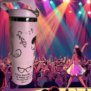May include: A light pink tumbler with a white handle and silver accents. The tumbler features black graphics of a cat, musical notes, and a woman's silhouette, along with the word "Swiftie" and a quote. The background shows a stage with a performer and a crowd.