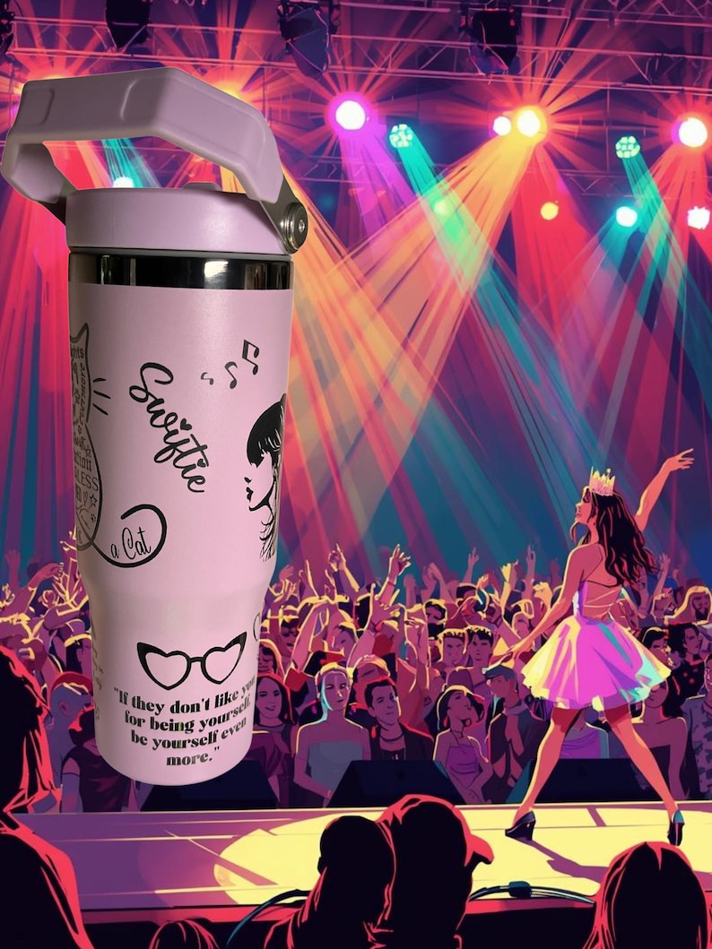 May include: A light pink tumbler with a white handle and silver accents. The tumbler features black graphics of a cat, musical notes, and a woman's silhouette, along with the word "Swiftie" and a quote. The background shows a stage with a performer and a crowd.