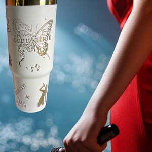 May include: A white tumbler with a gold rim and handle, featuring a butterfly design and the word "reputation". The tumbler also has musical notes and a silhouette of a person. The person is holding a black microphone.
