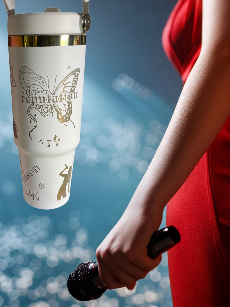 May include: A white tumbler with a gold rim and handle, featuring a butterfly design and the word "reputation". The tumbler also has musical notes and a silhouette of a person. The person is holding a black microphone.