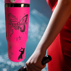 May include: A bright pink tumbler with a black butterfly and snake design, the word "reputation", musical notes, and a silhouette of a singer. The tumbler has a handle and a black lid.