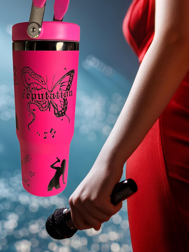 May include: A bright pink tumbler with a black butterfly and snake design, the word "reputation", musical notes, and a silhouette of a singer. The tumbler has a handle and a black lid.