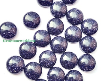 May include: A collection of round, polished blue goldstone cabochons with sparkling inclusions. The gemstones are scattered on a white surface, showcasing their deep blue color and reflective qualities. These stones are ideal for jewelry making.