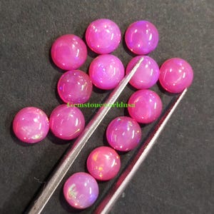 Puede incluir: Una colección de piedras preciosas rosas, redondas y pulidas. Las piedras preciosas son de un color magenta vibrante y parecen tener una superficie lisa y brillante. Las piedras son sujetadas por unas pinzas plateadas sobre un fondo oscuro.