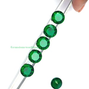 May include: A collection of round, faceted emerald green gemstones. The gems are arranged in a diagonal line, held by a silver tool, with one gem separate. The image includes the text "Gemstoneworkus".