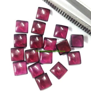 May include: A collection of 16 square-shaped, faceted, red gemstones. The gemstones are loose and scattered on a white surface.