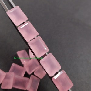 Könnte beinhalten: Eine Gruppe von rechteckigen rosa Edelsteinen, einige befinden sich auf einer Metallpinzette, andere sind auf einer grauen Oberfläche verstreut. Gemstoneworldusa ist in Grün auf dem Bild gedruckt.