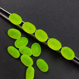 Op de afbeelding: Een verzameling ovaalvormige, neon groene edelstenen. Sommige worden vastgehouden door een zilveren pincet, terwijl andere verspreid liggen op een donkergrijze ondergrond. De edelstenen hebben een doorschijnende kwaliteit.