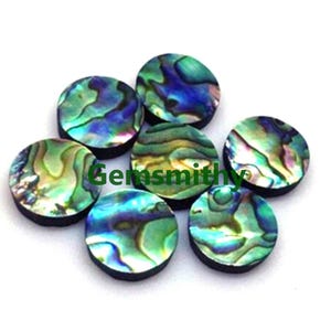 May include: Seven round abalone shell cabochons with iridescent blue, green, and gold hues. The cabochons have a dark rim and are scattered on a white surface. The text "Gemsmithy" is visible in green.