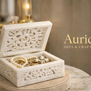 Può includere: Un portagioie in marmo bianco con intricate incisioni floreali, aperto per rivelare una collezione di gioielli, tra cui anelli, collane e bracciali. La scatola è esposta su un tavolo rotondo, con il testo "Auric ARTS & CRAFTS" in oro.