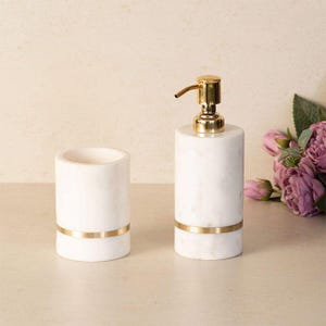 May include: A white marble soap dispenser and tumbler set. The cylindrical dispenser has a gold-colored pump and a gold band. The tumbler is a matching white marble with a gold band. The set is designed for bathroom use.