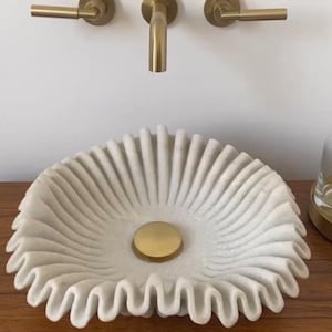 Può includere: Un lavabo in marmo bianco con un bordo arricciato e uno scarico dorato. Il lavabo è montato su un piano di lavoro in legno.