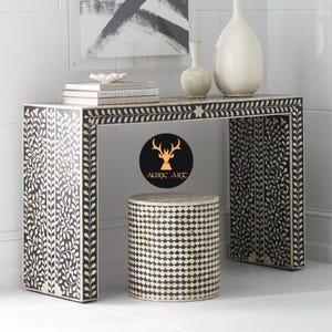 May include: A black and white decorative console table and matching stool. The table and stool feature a leaf and geometric pattern. The table has a rectangular top with a leaf design border. The stool is cylindrical with a geometric pattern. The table has two vases and books on top.