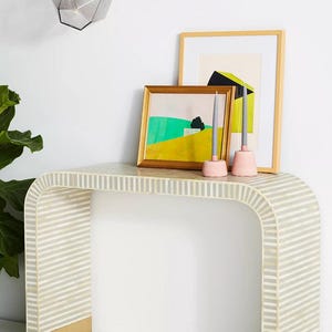 May include: A modern console table with a striped pattern, gold accents, and a pair of coral-colored flats. The table displays framed artwork and pink candle holders with gray candles. A geometric pendant light hangs above.