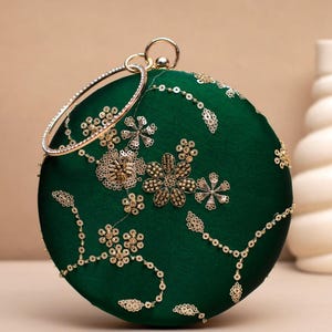 Peut inclure: Une pochette ronde vert émeraude avec une poignée dorée. La pochette est ornée de sequins dorés et de motifs floraux. La poignée est un anneau doré avec une rangée de strass transparents. Un accessoire de soirée.