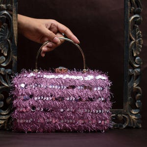 Puede incluir: Un bolso de mano malva con un exterior texturizado y con flecos y lentejuelas plateadas. El bolso rectangular tiene un asa dorada y un cierre decorativo. El bolso es sostenido por una mano, lo que sugiere su tamaño y portabilidad.