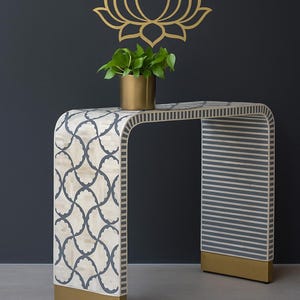 Op de afbeelding: Een decoratieve consoletafel met een gebogen ontwerp. De tafel heeft een crèmekleurig oppervlak met een herhalend blauw patroon aan één kant en verticale strepen aan de andere kant. De basis en poten zijn goudkleurig. Een gouden lotusbloem staat op de muur boven de tafel.