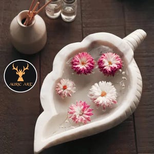 May include: White marble leaf-shaped bowl with a handle, filled with water and pink and white flowers.